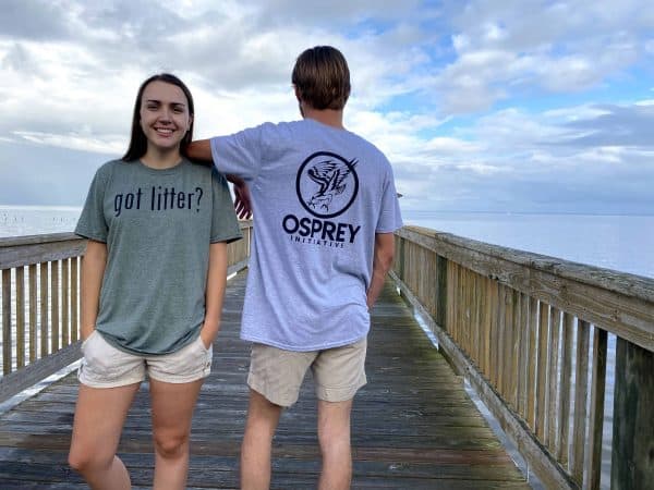 A male and female are standing on a wooden pier. The female is smiling at the camera and wearing a green “Got Litter” t-shirt. The male is facing away from the camera and wearing a grey “Got Litter?” t-shirt.