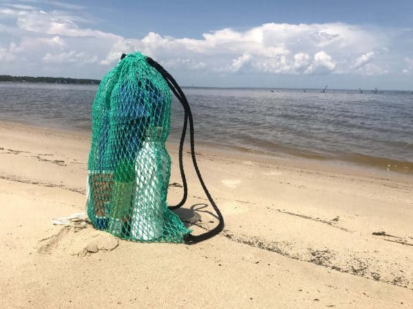 The Osprey Litter Gitter Backpack is filled with beach items and sitting on the beach. The bag features green mesh with black handles and a cinch top to close it.