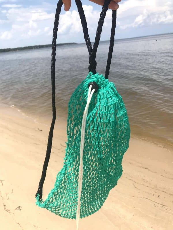 The Osprey Litter Gitter Backpack is empty and being held up with the beach and water in the background. The bag features green mesh with black handles and a cinch top to close it.