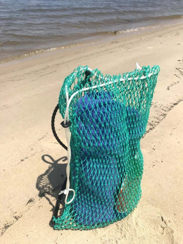 The Osprey Litter Gitter Backpack is filled with beach items and sitting on the beach. The bag features green mesh with black handles and a cinch top to close it.