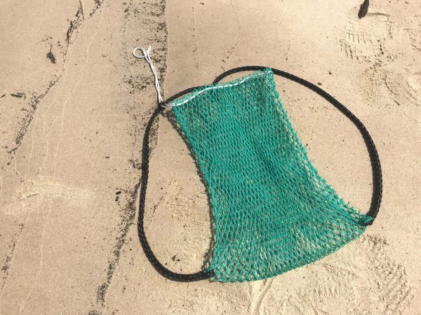 The Osprey Litter Gitter Backpack is empty and laying flat on the beach. The bag features green mesh with black handles and a cinch top to close it.