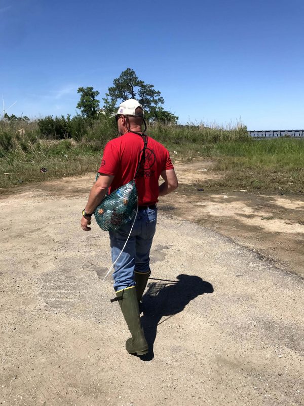 A man wearing a red shirt walks while wearing the Litter Gitter bag on his back. He is walking on a paved surface, toward a grassy area to collect litter.