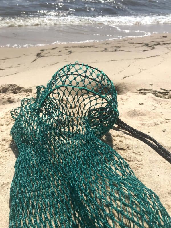 The empty Osprey Litter Gitter bag is being held with the beach in the background. It is a bag made of green mesh material with black handles and a cinch bottom, to allow to release litter. It has an opening that stays open so you can easily put litter in it while walking or jogging.