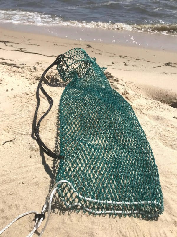 The empty Osprey Litter Gitter bag is being held with the beach in the background. It is a bag made of green mesh material with black handles and a cinch bottom, to allow to release litter. It has an opening that stays open so you can easily put litter in it while walking or jogging.