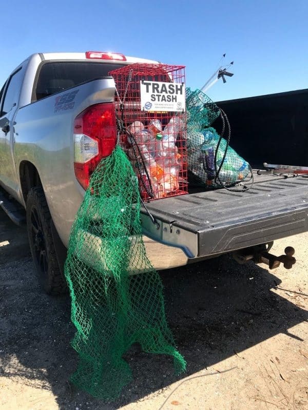 The Osprey Reusable Recycle bag is shown hanging on the back tailgate of a pick up truck. It is also shown with the Osprey Trash Stash in the pick truck bed. The bag is made of a green mesh material with black handles that cinch the top shut.