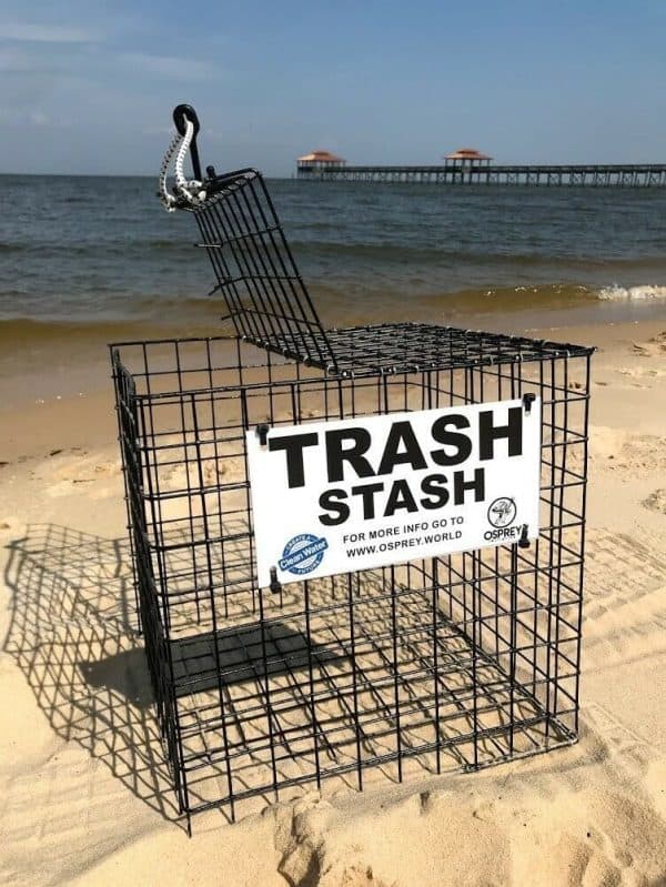 The Trash Stash is pictured on the beach, in the color black in the cube size. It is made of rubber coated crab trap wire and has a sign that says "Trash Stash" in black and white with the Osprey logo. It has a top that opens and secures shut with a bungee cord.