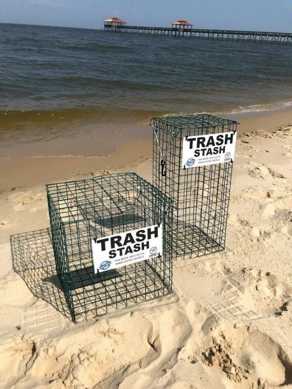 The Trash Stash is pictured on the beach, in the color green and in two sizes (cube and tower). It is made of rubber coated crab trap wire and has a sign that says "Trash Stash" in black and white with the Osprey logo. It has a top that opens and secures shut with a bungee cord.