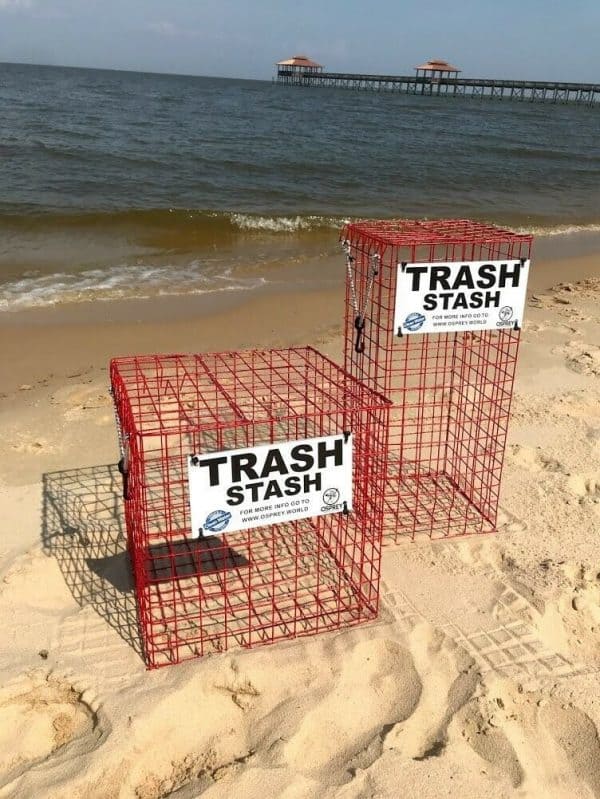 The Trash Stash is pictured on the beach, in the color red and in two sizes (cube and tower). It is made of rubber coated crab trap wire and has a sign that says "Trash Stash" in black and white with the Osprey logo. It has a top that opens and secures shut with a bungee cord.