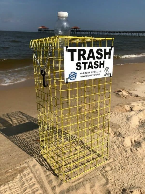 The Trash Stash is pictured on the beach, in the color yellow and in the tower size. It is made of rubber coated crab trap wire and has a sign that says "Trash Stash" in black and white with the Osprey logo. It has a top that opens and secures shut with a bungee cord. A water bottle is being placed inside the Trash Stash.