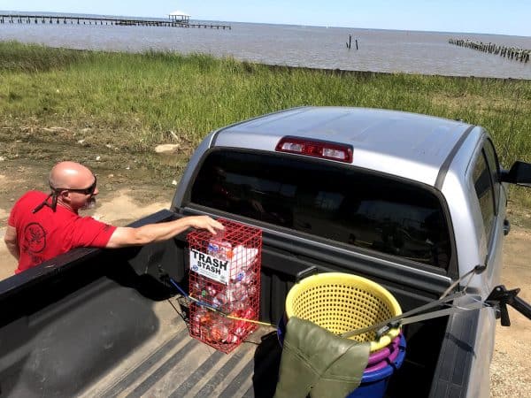 A man uses the red tower Trash Stash in the back of his pickup truck bed. He is placed an empty water bottle inside the Trash Stash while enjoying the outdoors on Mobile Bay.