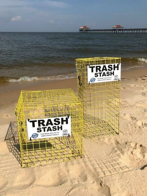 The Trash Stash is pictured on the beach, in the color yellow and in two sizes (cube and tower). It is made of rubber coated crab trap wire and has a sign that says "Trash Stash" in black and white with the Osprey logo. It has a top that opens and secures shut with a bungee cord.