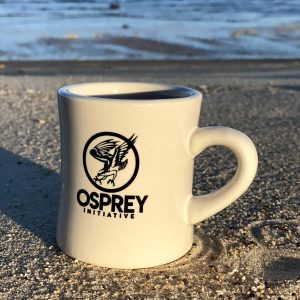 A white coffee mug with the black Osprey logo on it is on the beach.