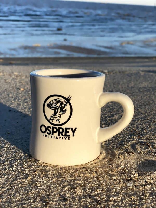 A white coffee mug with the black Osprey logo on it is on the beach.