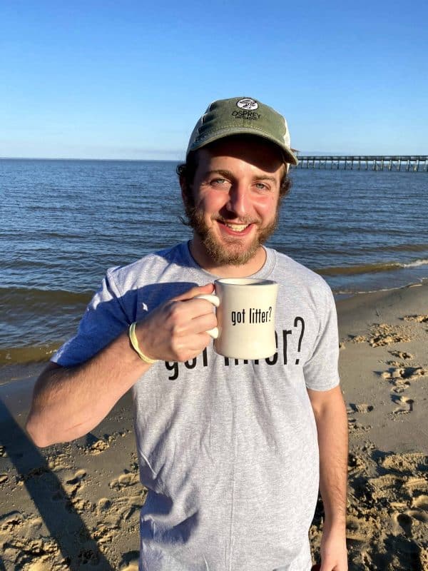 A smiling male is holding the Osprey coffee mug, showing the side that says "got litter?" in black text. He is on the beach and wearing an Osprey "Got Litter?" t-shirt and Osprey baseball hat.