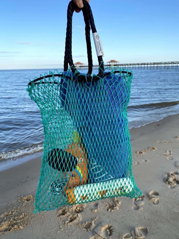 The Osprey tote bag is being held with the beach in the background. The bag contains beach items, including sunscreen and a towel. The bag is made of green mesh material with black rope handles.