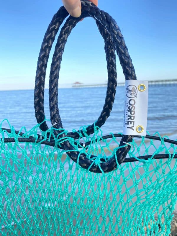 A close up photo of the Osprey tote bag black handles, with the Osprey logo tag on them. The green mesh of the bag is also visible and the beach and water are in the background.