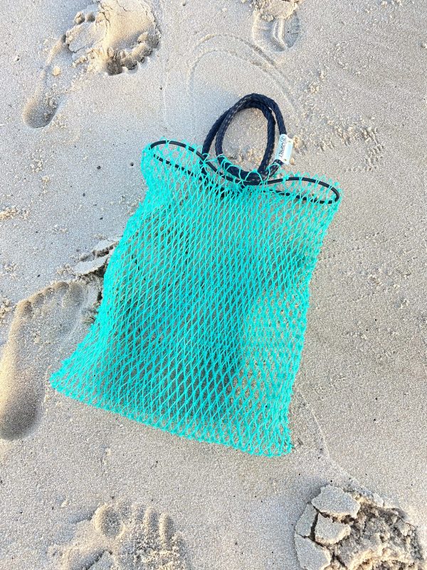 An empty Osprey tote bag is laid on the sandy beach. The bag is made of green mesh and has large black rope handles.