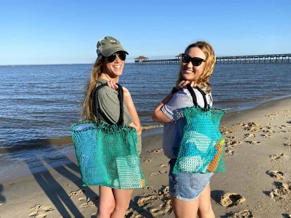 Two females are holding the Osprey tote bags on the beach. Their bags contain beach items, including sunscreen and towels. They are also wearing Osprey t-shirts and hats.