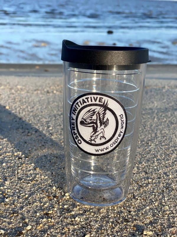 A clear tervis tumbler is positioned on the beach. The tumbler features a black and white Osprey logo with a black lid.
